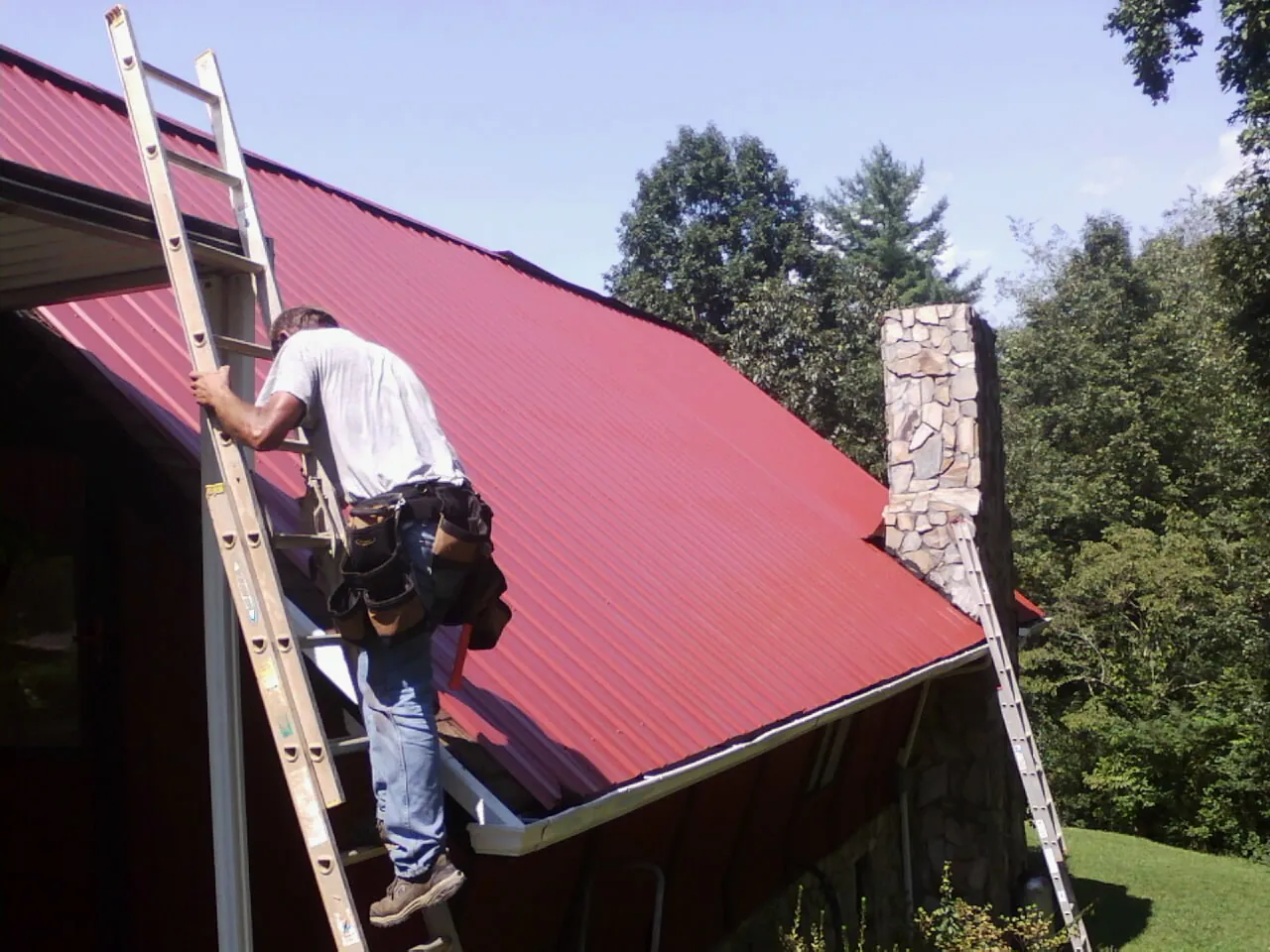 Roofer on a ladder beside a red metal roof and stone chimney — Soffit & Fascia Repair by Roofing Brothers in Rowlesburg